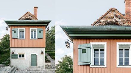 Ein kleines, freistehendes Haus mit rechteckigem Grundriss steht am Hang in einer grünen Umgebung, der untere Teil ist verputzt und massiv, der obere Teil besteht aus vertikal verschalten Holzflächen in warmem Farbton, grüne Fensterläden, ein steiles Ziegeldach und ein gemauerter Schornstein prägen das Erscheinungsbild, eine Außentreppe führt seitlich nach oben, wodurch das Bild ein sorgfältig instand gesetztes oder neu interpretiertes Weinberg- oder Gartenhaus zeigt, das traditionelle Bauweise mit zeitgemäßer Nutzung verbindet und für den behutsamen Umgang mit kleinteiliger historischer Architektur und Landschaft steht.