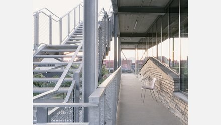 Außenumlauf mit Glasfassade rechts und Ziegelbrüstung; links Stahl-Außentreppe; einzelner Stuhl, warmes Abendlicht.