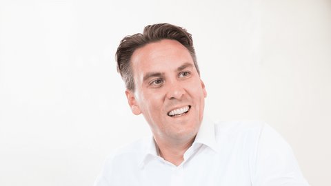 Porträt einer lächelnden Person mit kurzem, dunklem Haar in weißem Hemd; Blick nach rechts vor hellem Hintergrund.
