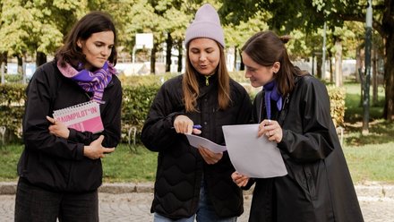 Das Bild zeigt drei Personen, die im Freien auf einem gepflasterten Weg stehen, umgeben von Bäumen und Grünflächen. Sie tragen wetterfeste Kleidung und halten Notizblöcke und lose Blätter in den Händen. Eine Person zeigt mit einem Stift auf ein Dokument, während die anderen aufmerksam mitlesen. Die Situation wirkt konzentriert und kooperativ, als würden sie gemeinsam etwas planen oder überprüfen. Zweck des Bildes ist es, partizipative Arbeit im öffentlichen Raum zu zeigen, etwa gemeinsames Planen, Diskutieren oder Bewerten von Projekten vor Ort.