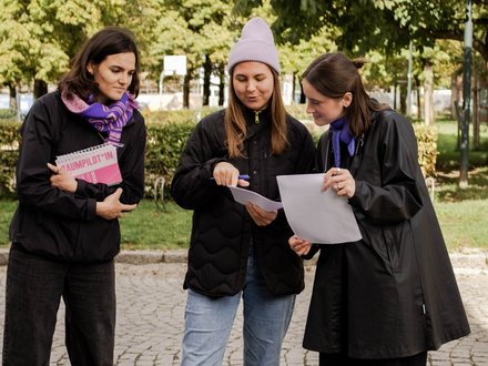 Das Bild zeigt drei Personen, die im Freien auf einem gepflasterten Weg stehen, umgeben von Bäumen und Grünflächen. Sie tragen wetterfeste Kleidung und halten Notizblöcke und lose Blätter in den Händen. Eine Person zeigt mit einem Stift auf ein Dokument, während die anderen aufmerksam mitlesen. Die Situation wirkt konzentriert und kooperativ, als würden sie gemeinsam etwas planen oder überprüfen. Zweck des Bildes ist es, partizipative Arbeit im öffentlichen Raum zu zeigen, etwa gemeinsames Planen, Diskutieren oder Bewerten von Projekten vor Ort.