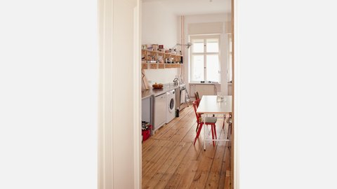 "Blick durch eine offene helle Türe in eine moderne Wohnküche mit Holzparkett. Die Küche verfügt über weiße Geräte, eine Waschmaschine, eine Spüle mit Fenster und hölzerne Regale an der Wand. Ein weißer Esstisch mit roten Stühlen steht im Wohnbereich."