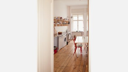 "Blick durch eine offene helle Türe in eine moderne Wohnküche mit Holzparkett. Die Küche verfügt über weiße Geräte, eine Waschmaschine, eine Spüle mit Fenster und hölzerne Regale an der Wand. Ein weißer Esstisch mit roten Stühlen steht im Wohnbereich."