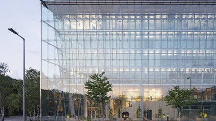 Transparente Gebäudehülle mit Blick in einen beleuchteten Innenraum.