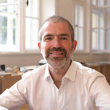 Porträt eines Mannes in hellem Innenraum, sitzend auf einem roten Sofa vor großen Fenstern.