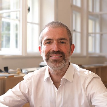 Porträt eines Mannes in hellem Innenraum, sitzend auf einem roten Sofa vor großen Fenstern.