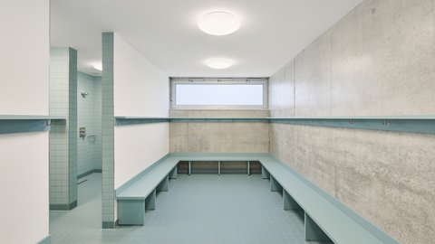 Leerer Umkleideraum mit hellblauen Sitzbänken entlang der Wände, grauem Boden und Betonwand mit schmalem Fenster