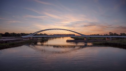 Eisenbahnbrücke mit Stahlbogen bei Sonnenuntergang, ein Zug fährt über den Fluss, Himmel spiegelt sich im Wasser.