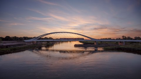 Eisenbahnbrücke mit Stahlbogen bei Sonnenuntergang, ein Zug fährt über den Fluss, Himmel spiegelt sich im Wasser.
