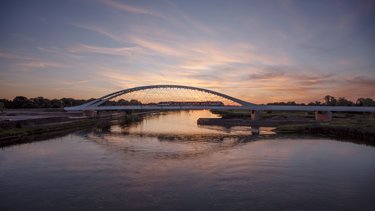 Eisenbahnbrücke mit Stahlbogen bei Sonnenuntergang, ein Zug fährt über den Fluss, Himmel spiegelt sich im Wasser.