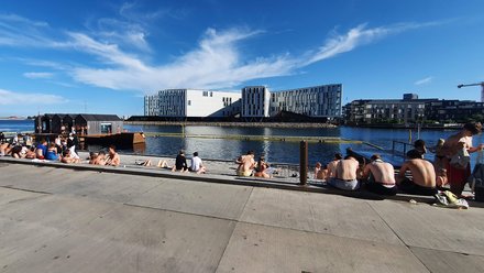 Menschen entspannen an einem sonnigen Hafenbecken auf einer breiten Uferpromenade. Einige sitzen am Wasser, andere schwimmen oder genießen den Blick auf moderne Gebäude.