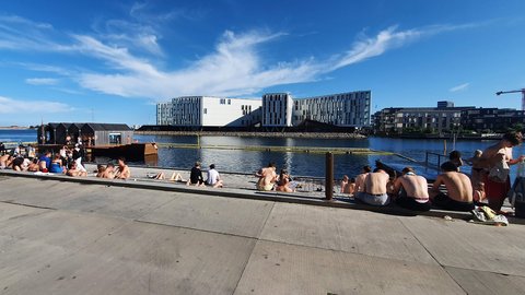 Menschen entspannen an einem sonnigen Hafenbecken auf einer breiten Uferpromenade. Einige sitzen am Wasser, andere schwimmen oder genießen den Blick auf moderne Gebäude.
