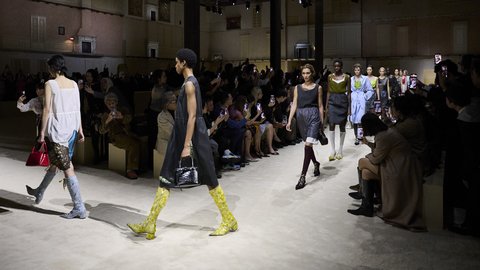 "Modenschau in einer großen historischen Halle mit schwarzem Industriedachgerüst. Models präsentieren zeitgenössische Mode auf einem beleuchteten, ungepflasterten Laufsteg. Im Vordergrund läuft ein Model in weißem Top und auffälligen neon-gelben Stiefeln. Weitere Models folgen in verschiedenen Outfits. Zuschauer sitzen beidseitig des Laufstegs und fotografieren mit Smartphones. Die Halle hat mehrere Säulen und historische Architekturelemente. Kunstvolles Staging durch Lichtsetzung und Raumaufteilung."