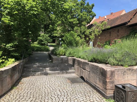 Schmaler gepflasterter Fußweg mit Steinstufen. Links und rechts Steinmauern. Dichte grüne Vegetation und Lavendelstrauch mit lila Blüten rechts. Schattig durch überhängende Bäume. Historisches Ziegelgebäude mit Dach sichtbar. Ruhige, ländliche Atmosphäre.