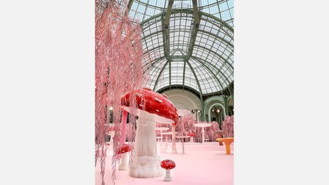 "Detailansicht einer künstlerischen Installation mit großen, rot-weißen Pilzen und rosafarbenen, hängenden Elementen. Die Installation steht unter einer hohen, gewölbten Glasdecke mit sichtbarer Stahlstruktur und wirkt wie eine künstliche Landschaft."