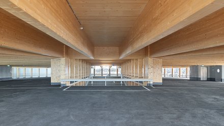 Innenansicht eines Holzparkhauses, sichtbare Holzdecken und -stützen, offene Ebenen mit Fahrbahnmarkierungen.