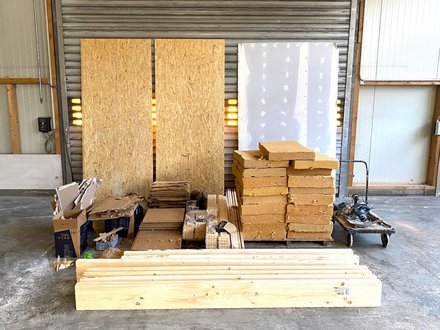 Sortierte Baustoffe in einer Halle: Bretter, OSB‑ und Gipsplatten, stapelweise Dämmblöcke auf Palette, Kartons und Transportwagen.