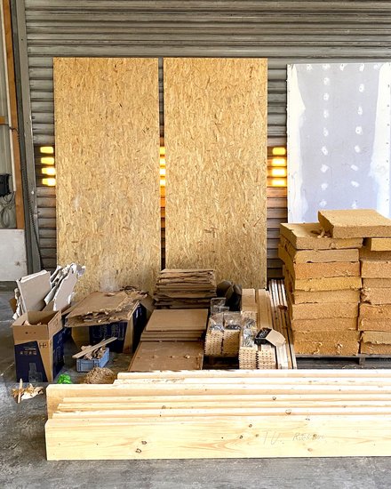 Sortierte Baustoffe in einer Halle: Bretter, OSB‑ und Gipsplatten, stapelweise Dämmblöcke auf Palette, Kartons und Transportwagen.