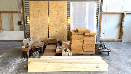 Sortierte Baustoffe in einer Halle: Bretter, OSB‑ und Gipsplatten, stapelweise Dämmblöcke auf Palette, Kartons und Transportwagen.