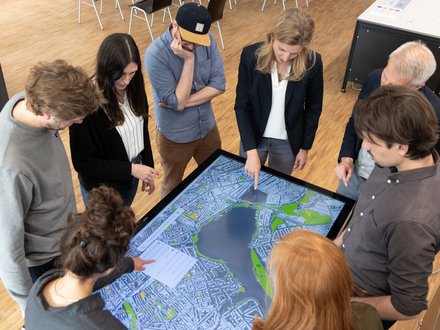 Acht Personen stehen um einen großen, liegenden Touchscreen mit Stadtplan; sie zeigen auf die Karte und diskutieren die Planung.