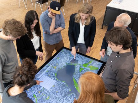 Acht Personen stehen um einen großen, liegenden Touchscreen mit Stadtplan; sie zeigen auf die Karte und diskutieren die Planung.