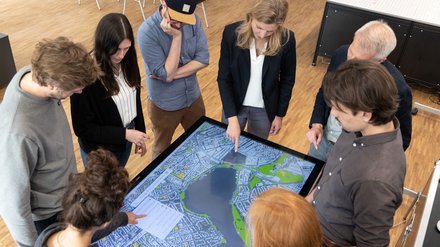 Acht Personen stehen um einen großen, liegenden Touchscreen mit Stadtplan; sie zeigen auf die Karte und diskutieren die Planung.