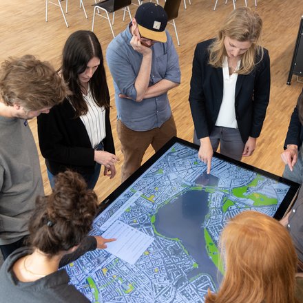 Acht Personen stehen um einen großen, liegenden Touchscreen mit Stadtplan; sie zeigen auf die Karte und diskutieren die Planung.