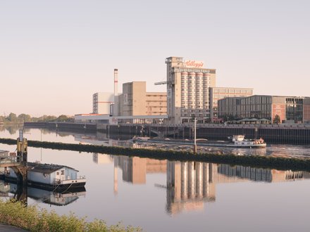 Ruhiger Fluss im weichen Morgenlicht; am rechten Ufer Industrie- und Speicherbauten mit hohem Silo und „Kellogg’s“-Schriftzug. Ein kleines weißes Schiff fährt vor der Kaimauer, links liegt ein Hausboot; die Gebäude spiegeln sich im Wasser.