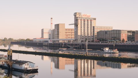 Ruhiger Fluss im weichen Morgenlicht; am rechten Ufer Industrie- und Speicherbauten mit hohem Silo und „Kellogg’s“-Schriftzug. Ein kleines weißes Schiff fährt vor der Kaimauer, links liegt ein Hausboot; die Gebäude spiegeln sich im Wasser.