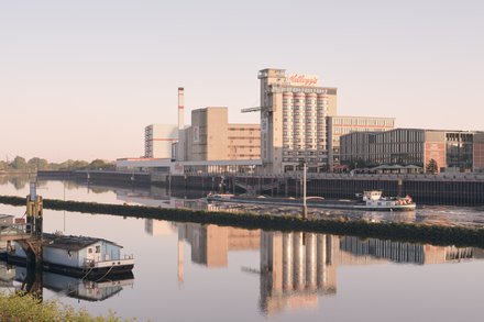 Ruhiger Fluss im weichen Morgenlicht; am rechten Ufer Industrie- und Speicherbauten mit hohem Silo und „Kellogg’s“-Schriftzug. Ein kleines weißes Schiff fährt vor der Kaimauer, links liegt ein Hausboot; die Gebäude spiegeln sich im Wasser.