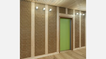 Flurdetail: Lehmziegel-Felder zwischen Holzständern; zentrale, grüne Tür, Wandstrahler an der Decke.