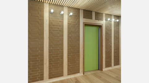 Flurdetail: Lehmziegel-Felder zwischen Holzständern; zentrale, grüne Tür, Wandstrahler an der Decke.