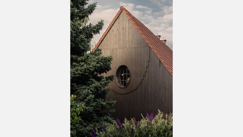 Nahaufnahme der Giebelseite desselben Hauses wie in image_31.png, aber aus einem anderen Winkel. Die braune Holzverkleidung und das rote Ziegeldach sind deutlich zu sehen. Ein einzelnes rundes Fenster mit schwarzem Rahmen befindet sich auf der Giebelebene. Links vom Haus steht eine große, dichte grüne Tanne. Im Vordergrund blühen lila Schmetterlingsflieder und andere grüne Pflanzen. Der Himmel ist grau-blau und leicht bewölkt.