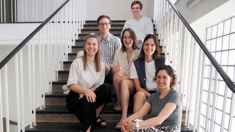 Das Bild zeigt eine Gruppe von sieben jungen Erwachsenen, die auf einer breiten Treppe in einem hellen Innenraum sitzen. Sie schauen in die Kamera und lächeln, die Stimmung wirkt offen und kollegial. Die Umgebung ist schlicht, mit weißen Geländern und viel Tageslicht. Die Personen sitzen locker verteilt auf den Stufen, was Nähe und Teamgeist vermittelt. Zweck des Bildes ist es, ein Team oder eine Projektgruppe sichtbar zu machen und Zusammenarbeit sowie Vielfalt innerhalb einer Organisation oder Initiative zu zeigen.