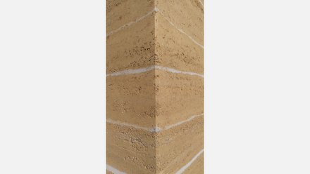 Detail: scharfe Außenecke einer Stampflehmwand mit hellen horizontalen Schichten und rauer Textur.