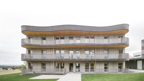 Mehrgeschossiges Wohngebäude mit geschwungenen Balkonen und vertikaler Holzlattung, davor Rasenfläche und gepflasterter Weg.