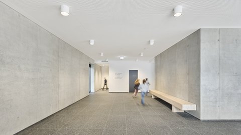Heller, moderner Flur mit Betonwänden, Sitzbank an der rechten Wand und zwei laufenden Kindern