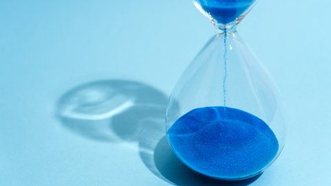 „Eine gläserne Sanduhr auf hellblauem Hintergrund, in der leuchtend blauer Sand nach unten rieselt.“