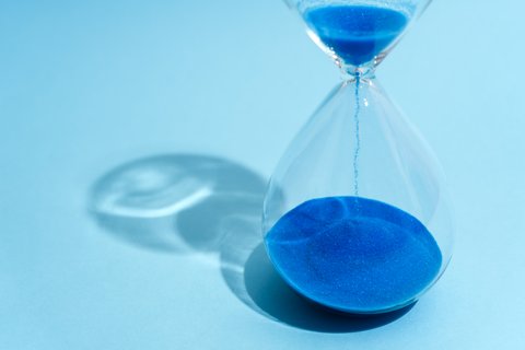 „Eine gläserne Sanduhr auf hellblauem Hintergrund, in der leuchtend blauer Sand nach unten rieselt.“