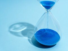 „Eine gläserne Sanduhr auf hellblauem Hintergrund, in der leuchtend blauer Sand nach unten rieselt.“