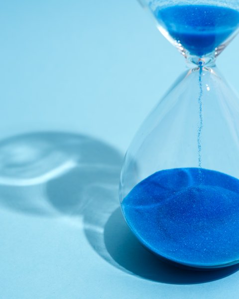 „Eine gläserne Sanduhr auf hellblauem Hintergrund, in der leuchtend blauer Sand nach unten rieselt.“