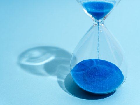 „Eine gläserne Sanduhr auf hellblauem Hintergrund, in der leuchtend blauer Sand nach unten rieselt.“