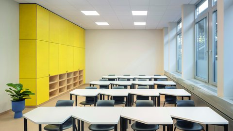 "Moderner Schulraum mit klarem Design. Links eine große gelbe Schrankelement-Wand mit integrierten Holzfächern darunter. Mehrere weiße trapezförmige Schülertische mit schwarzen Kunststoffstühlen in Reihen angeordnet. Fensterfront rechts mit Blick nach außen. Weiße Wände, heller Holzboden, weiße Decke mit quadratischer Beleuchtung. Eine grüne Topfpflanze."