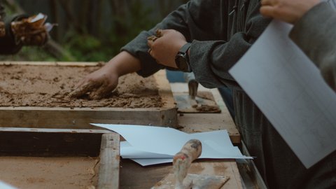 Personen arbeiten an einer Lehmstation und formen feuchten Lehm in Holzrahmen. Auf dem Arbeitstisch liegen Maurerkellen und Unterlagen, während Hände den Lehm glätten und bearbeiten.