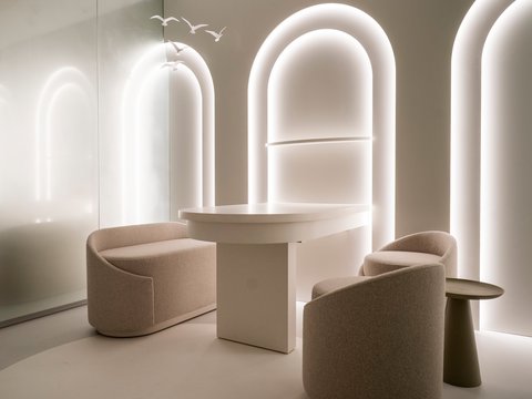 "Ähnliche Ansicht eines modernen Loungebereichs aus leicht anderer Perspektive. Ein niedriger beige-weißer zylindrischer Tisch steht zentral, flankiert von zwei ovalen Sesseln in Beige. Rechts ein konischer Beistelltisch. Dahinter drei große bogenförmige beleuchtete Fenster. Der Raum wirkt luftig und minimalistisch in hellen Farbtönen."