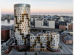 Gesamtansicht des Ensembles in städtischer Situation: weiträumige Außenaufnahme bei Dämmerung zeigt den runden Turm und die niedrigeren, organisch gegliederten Baukörper davor, Hafenanlagen am Horizont; Straße und Nebengebäude im Vordergrund.