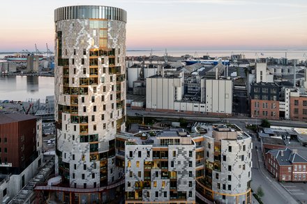 Gesamtansicht des Ensembles in städtischer Situation: weiträumige Außenaufnahme bei Dämmerung zeigt den runden Turm und die niedrigeren, organisch gegliederten Baukörper davor, Hafenanlagen am Horizont; Straße und Nebengebäude im Vordergrund.