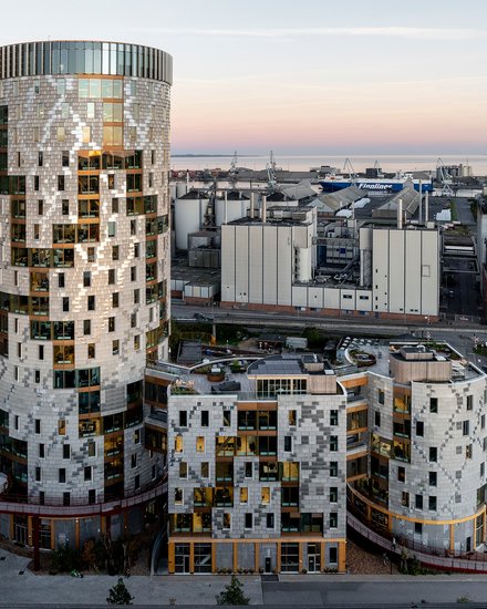 Gesamtansicht des Ensembles in städtischer Situation: weiträumige Außenaufnahme bei Dämmerung zeigt den runden Turm und die niedrigeren, organisch gegliederten Baukörper davor, Hafenanlagen am Horizont; Straße und Nebengebäude im Vordergrund.