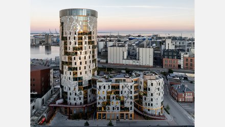 Gesamtansicht des Ensembles in städtischer Situation: weiträumige Außenaufnahme bei Dämmerung zeigt den runden Turm und die niedrigeren, organisch gegliederten Baukörper davor, Hafenanlagen am Horizont; Straße und Nebengebäude im Vordergrund.