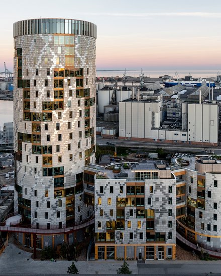 Gesamtansicht des Ensembles in städtischer Situation: weiträumige Außenaufnahme bei Dämmerung zeigt den runden Turm und die niedrigeren, organisch gegliederten Baukörper davor, Hafenanlagen am Horizont; Straße und Nebengebäude im Vordergrund.